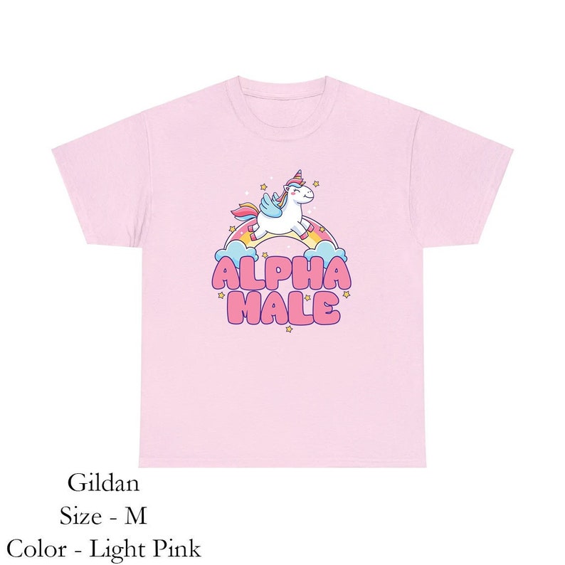 Puede incluir: Una camiseta rosa claro con un unicornio de dibujos animados montando un arco&iacute;ris y el texto "ALPHA MALE" en letras rosas.