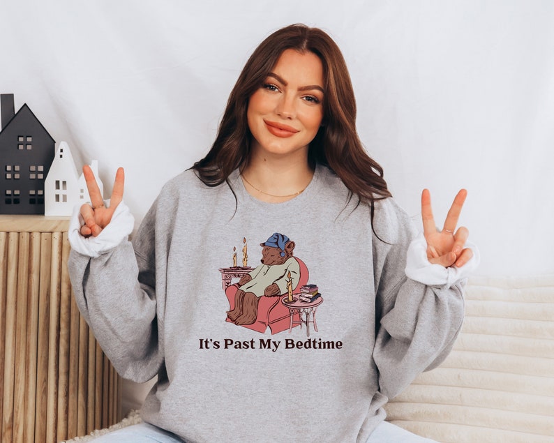 May include: A gray sweatshirt with a cartoon illustration of a bear wearing a nightcap and pajamas, sitting in a chair with a book and a lit candle. The text "It's Past My Bedtime" is printed below the illustration.