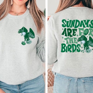 Op de afbeelding: Grijs sweatshirt met een groene adelaar afbeelding op de voorkant en de tekst "Sundays are for the birds" op de achterkant. De adelaar houdt een voetbal vast.