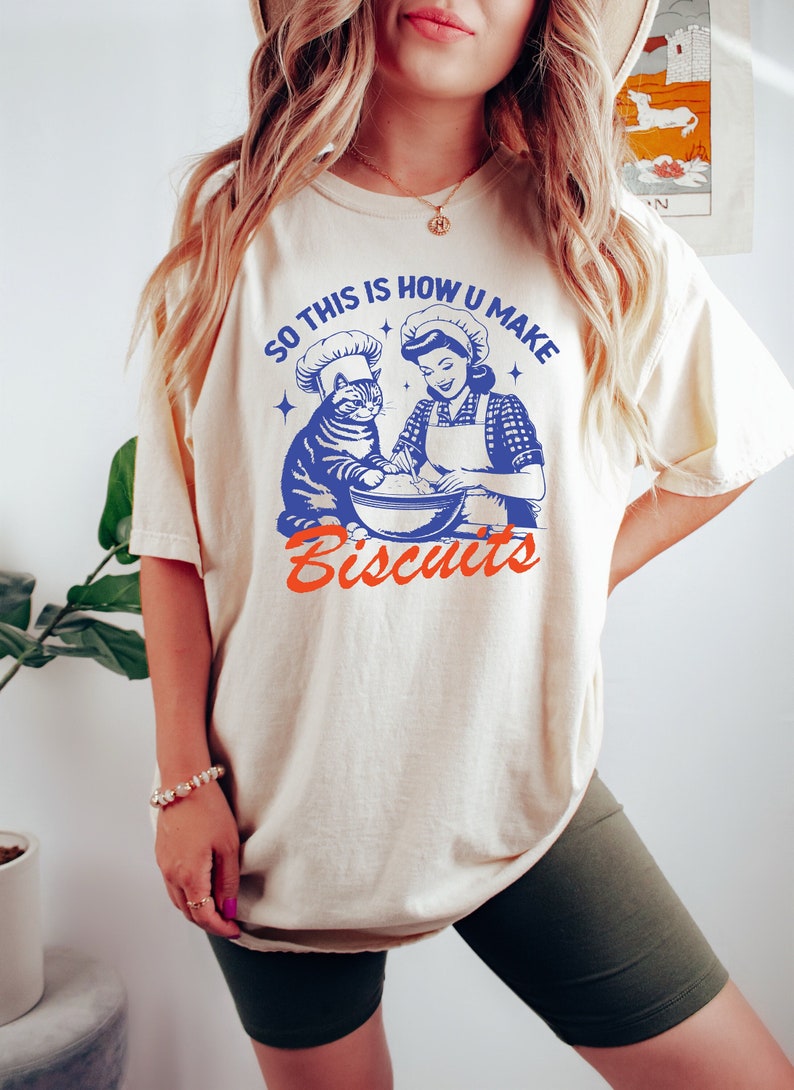 May include: Cream-colored t-shirt with a blue and orange graphic. The graphic features a cat and a woman in chef hats, mixing ingredients in a bowl. Text reads "SO THIS IS HOW U MAKE Biscuits."