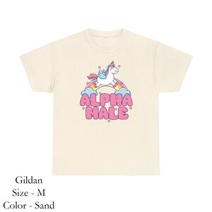 Puede incluir: Una camiseta de color arena con un unicornio de dibujos animados montando un arco&iacute;ris y el texto "ALPHA MALE" en letras rosas.