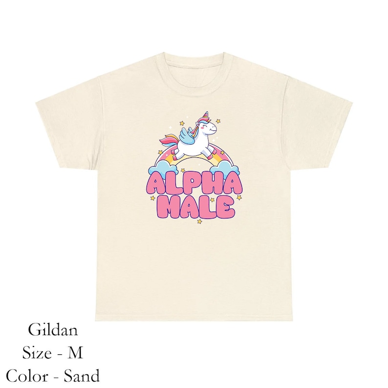 Puede incluir: Una camiseta de color arena con un unicornio de dibujos animados montando un arco&iacute;ris y el texto "ALPHA MALE" en letras rosas.