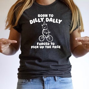 May include: A dark gray t-shirt with a white graphic of a bear riding a bicycle. The text on the shirt reads "Born to Dilly Dally, Forced to Pick Up the Pace."