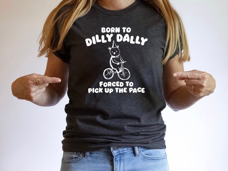 May include: A dark gray t-shirt with a white graphic of a bear riding a bicycle. The text on the shirt reads "Born to Dilly Dally, Forced to Pick Up the Pace."