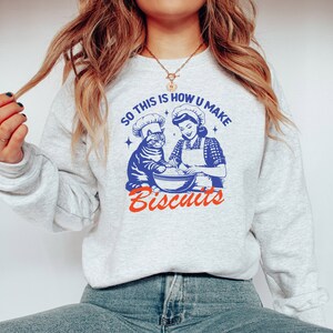 May include: A light gray sweatshirt with a retro-style graphic. The graphic features a cat and a woman in chef hats, mixing ingredients. Text reads "SO THIS IS HOW U MAKE Biscuits" in blue and red.