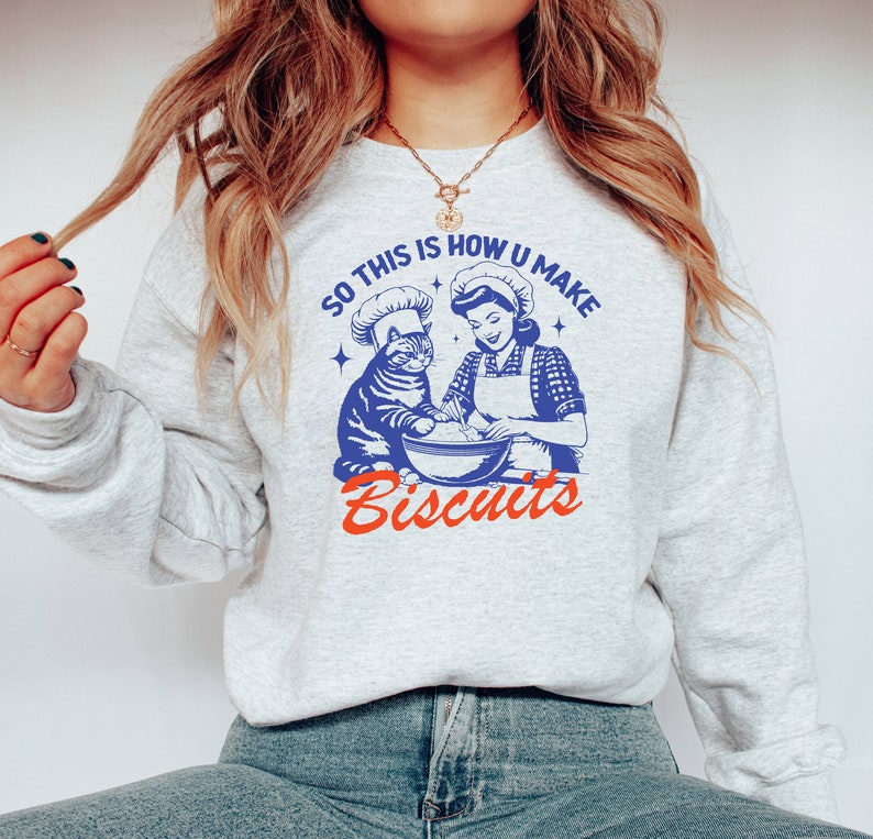 May include: A light gray sweatshirt with a retro-style graphic. The graphic features a cat and a woman in chef hats, mixing ingredients. Text reads "SO THIS IS HOW U MAKE Biscuits" in blue and red.