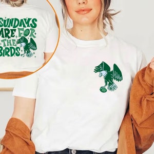 Op de afbeelding: Wit T-shirt met een groene adelaar afbeelding die een voetbal vasthoudt en de tekst "Sundays are for the Birds" op de achterkant.