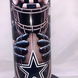 Puede incluir: Un vaso de acero inoxidable negro y azul con un gráfico de un jugador de fútbol americano con casco y sosteniendo un balón de fútbol. El balón de fútbol tiene una estrella blanca. El vaso tiene un efecto de goteo alrededor del gráfico.