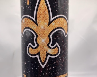 May include: A black tumbler featuring a gold fleur-de-lis emblem and the word "SAINTS" in gold. The design includes a starry background and gold dripping details.