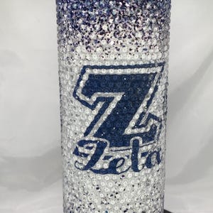 Può includere: Un bicchiere bianco e blu sfumato con un logo "7 Eleven" blu e bianco. Il bicchiere è ricoperto di strass.