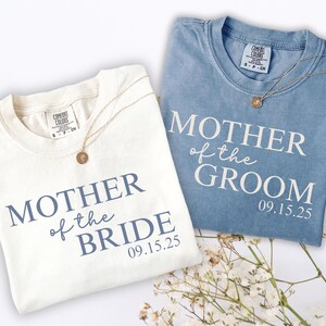 May include: A white t-shirt with blue text reading MOTHER of the BRIDE 09.15.25 and a blue t-shirt with white text reading MOTHER of the GROOM 09.15.25. Both shirts feature a Comfort Colors brand label at the inner neckline and a gold pendant necklace.