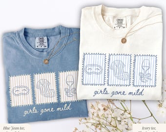 Puede incluir: Dos camisetas, una azul y otra marfil, con el texto "girls gone mild". Cada camiseta presenta tres ilustraciones enmarcadas: una máscara para dormir, un pijama y una copa de vino. Ambas camisetas tienen un collar dorado.