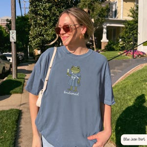 K&ouml;nnte beinhalten: Ein blau-graues Kurzarm-T-Shirt mit einer Grafik eines gr&uuml;nen Frosches, der ein blaues Kleid und eine wei&szlig;e Sch&auml;rpe mit der Aufschrift "Bridesmaid" tr&auml;gt. Der Frosch h&auml;lt ein Glas Champagner und das Wort "bridesmaid" steht darunter.