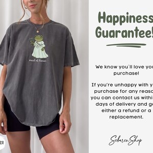 K&ouml;nnte beinhalten: Ein anthrazitfarbenes Kurzarm-T-Shirt mit einer Grafik eines Frosches in einem wei&szlig;en Kleid, der einen Blumenstrau&szlig; h&auml;lt, darunter der Text "maid of honor". Rechts steht der Text "Happiness Guarantee!" sowie Details zur R&uuml;ckgaberichtlinie.
