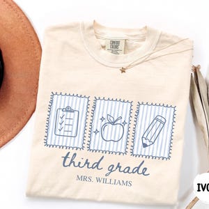 May include: An ivory short-sleeve t-shirt featuring a blue graphic of three rectangular frames containing a clipboard, an apple, and a pencil. Below the frames, the text reads third grade and MRS. WILLIAMS in a blue script and block font.