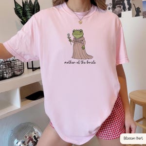 K&ouml;nnte beinhalten: Ein hellrosa T-Shirt mit einer Grafik eines gr&uuml;nen Frosches in einem langen altrosa Kleid mit kleiner Schleife, der eine wei&szlig;e Rose h&auml;lt. Der Frosch tr&auml;gt eine Sch&auml;rpe mit der Aufschrift Mother of the Bride und darunter steht mother of the bride.