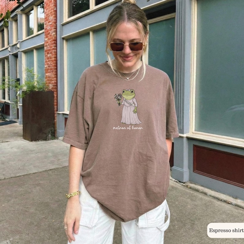 K&ouml;nnte beinhalten: Ein braunes Kurzarm-T-Shirt mit einer Grafik eines gr&uuml;nen Frosches, der ein hellrosa Kleid tr&auml;gt und einen Blumenstrau&szlig; h&auml;lt. Unter dem Frosch steht der Text "matron of honor" in einer wei&szlig;en Schreibschrift.