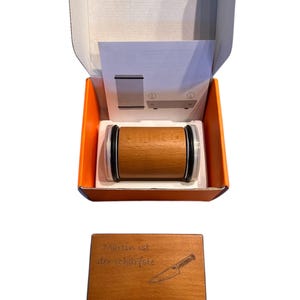 Könnte beinhalten: Ein Messer-Schärfer aus Holz mit zylindrischem Design, präsentiert in einer orange-weißen Box. Der Schärfer hat einen Holzkörper mit schwarzen Akzenten. Eine Holzplakette mit dem Text "Martin ist der schärfste" und einer Messerillustration ist ebenfalls enthalten.