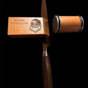 Könnte beinhalten: Ein Küchenmesser mit dunklem Holzgriff und Edelstahlklinge. Eine Holzbox mit dem Text "SCHARFE WEIHNACHTEN" und einer Weihnachtsmann-Grafik befindet sich neben dem Messer. Ein hölzerner Messerschärfer ist ebenfalls im Bild.