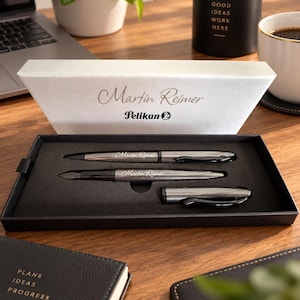 Könnte beinhalten: Ein Pelikan Stifteset in einer schwarzen Präsentationsbox mit weißem Deckel, der den Namen Martin Reimer trägt. Das Set enthält einen Kugelschreiber und einen Füllfederhalter, beide in metallisch grauer Optik mit eingraviertem Namen.