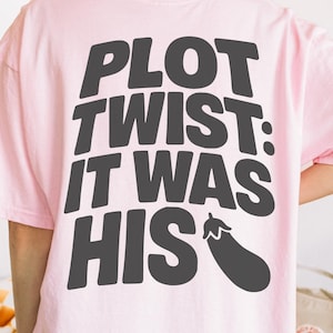 May include: Pale pink t-shirt with a black aubergine graphic and the text "Smut Slut" on the front. The back of the shirt features the text "PLOT TWIST: IT WAS HIS" with an aubergine graphic. A casual, unisex t-shirt.