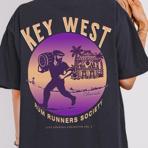 May include: Dark navy blue t-shirt with a circular graphic featuring a man carrying a barrel, the words "Key West" and "Rum Runners Society". The design has purple, orange, and yellow hues. The shirt has a relaxed fit.