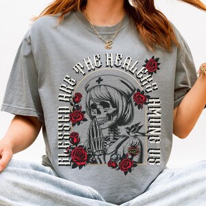 May include: Gray t-shirt with a graphic of a skeleton nurse surrounded by red roses and the text "BLESSED ARE THE HEALERS AMONG US". The shirt has short sleeves and a crew neck.