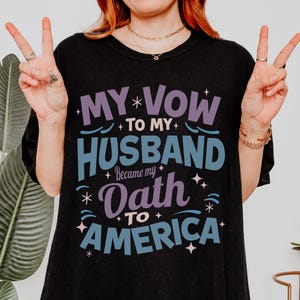 May include: Black t-shirt with the text "MY VOW TO MY HUSBAND Became my OATH TO AMERICA" in purple and blue. The shirt has a crew neck and short sleeves. The person in the image is giving a peace sign.