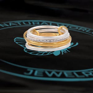 Puede incluir: Un conjunto de cuatro anillos apilables, dos plateados y dos dorados, con una fila de pequeñas piedras transparentes en el anillo superior.