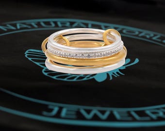 Könnte beinhalten: Ein Satz aus vier stapelbaren Ringen, zwei silber und zwei gold, mit einer Reihe kleiner, klarer Steine auf dem oberen Ring.