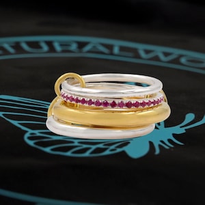 May include: A multi-band ring set with gold and silver bands, and a silver band with small red gemstones. A small gold ring connects the bands. The rings are on a black surface with a teal design.