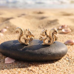 May include: A pair of gold-toned stud earrings shaped like squirrels, each holding a small acorn. The earrings feature a polished, reflective metallic finish with detailed engravings on the squirrel's body, tail, and face, showcasing a three-dimensional design.