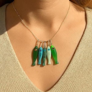 May include: A gold-toned necklace featuring six fish pendants in varying shades of green, blue, and white. Each fish has a small, colored eye. The necklace is worn against a neutral-toned top.