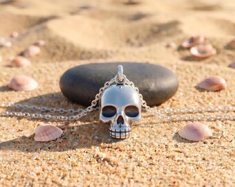 May include: A polished silver-toned skull pendant suspended from a matching delicate metal chain. The skull features detailed eye sockets, a nasal cavity, and defined teeth, resting against a smooth dark stone surface.