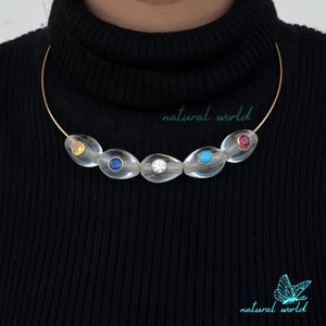 May include: A gold choker necklace with five clear oval-shaped beads, each with a different colored gemstone in the center. The gemstones are yellow, blue, clear, turquoise, and red.
