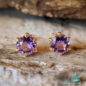 May include: A pair of gold stud earrings with round, purple amethyst gemstones. The earrings have a claw setting.