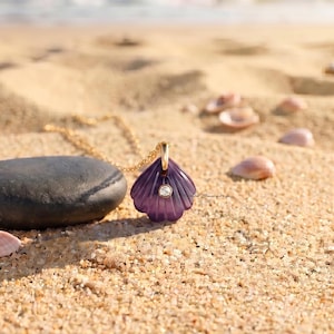 May include: A purple seashell-shaped pendant necklace featuring a small, clear, round gemstone set in the center. The pendant is suspended from a thin gold-toned chain and rests on a sandy surface next to a smooth, dark gray, rounded stone.