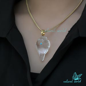 May include: A clear quartz crystal pendant with a turquoise stone in the center, hanging from a gold chain. The pendant is shaped like a seashell and has a delicate floral design.