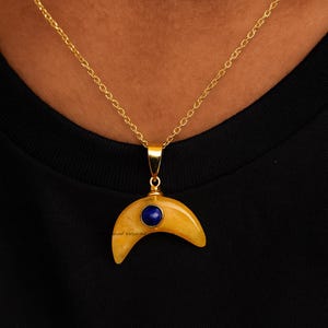 Puede incluir: Un collar dorado con un colgante en forma de media luna. El colgante es amarillo con una piedra preciosa azul en el centro. El collar se lleva sobre una camiseta negra.