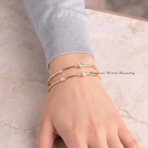 May include: Two gold-toned bangles featuring bezel-set round clear stones. The bracelets are worn on a wrist partially covered by a light grey long-sleeved garment. The text Natural World Jewelry is visible in the background.