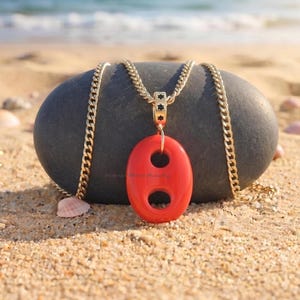 Può includere: Una collana a catena color oro con un ciondolo rosso liscio di forma ovale dotato di due aperture circolari. Il ciondolo è fissato alla catena tramite una contromaglia color oro decorata con piccole stelle nere. La catena è a maglia gourmette.