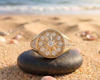 Puede incluir: Un anillo de oro con una cara circular que presenta un diseño de rosa de los vientos. El centro y las puntas de la estrella están engastados con pequeñas piedras redondas y transparentes. El aro y los lados también tienen incrustaciones.