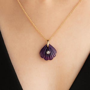 May include: A gold chain necklace with a pendant shaped like a purple seashell. The seashell pendant has a small, round, clear gemstone in the center. The necklace is worn against a person's skin and black clothing.
