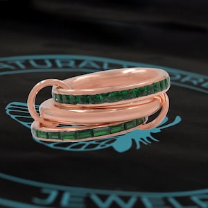 Könnte beinhalten: Drei ineinandergreifende Ringe aus Roségold mit einem Band aus quadratischen smaragdgrünen Edelsteinen um jeden Ring.