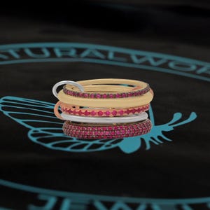 May include: A set of five stacking rings with a variety of colors and textures. The rings are made of metal and have a polished finish. The top ring is a thin, gold-colored band with a row of small, red gemstones. The second ring is a thicker, cream-colored band with a row of small, red gemstones. The third ring is a thin, rose gold-colored band with a row of small, red gemstones. The fourth ring is a thin, silver-colored band. The fifth ring is a thin, silver-colored band with a row of small, red gemstones.