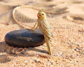 May include: A gold-toned necklace featuring a pendant shaped like a small bird with a light tan finish. The pendant is attached to a delicate gold-colored chain. The text Natural World Jewelry is visible on the bird's wing.