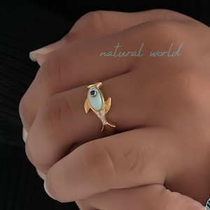 Può includere: Anello in oro con un design a forma di pesce blu e bianco con un solo occhio di pietra preziosa blu. L'anello è incastonato con piccole pietre preziose bianche attorno al corpo del pesce.