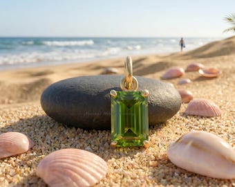 May include: A gold pendant featuring a rectangular emerald-cut green peridot gemstone held by four prongs. The pendant has a simple gold bail at the top and is positioned upright on a smooth dark gray stone, showcasing a vibrant green hue.