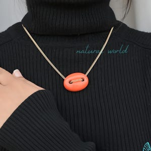 Puede incluir: Un collar de cadena dorada con un colgante grande de forma ovalada. El colgante es de color naranja brillante y tiene un acento dorado en el centro.