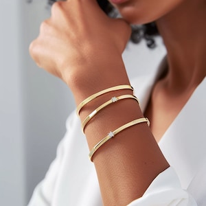 Peut inclure: Trois bracelets en or avec de petits accents de diamants. Les bracelets sont empilés sur le poignet d'une personne.